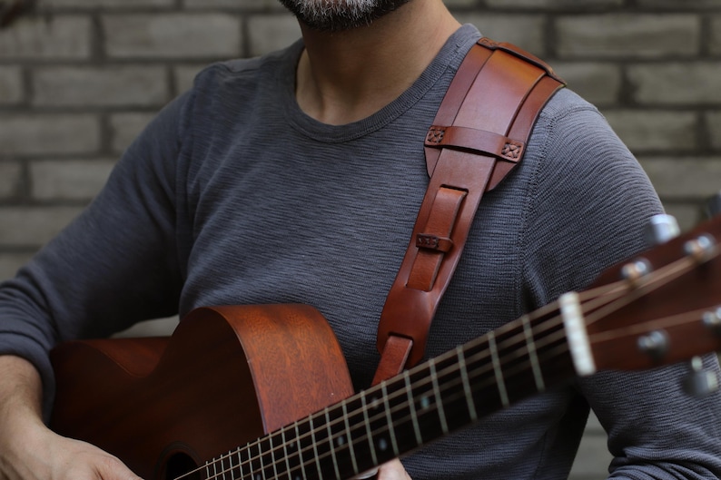 May include: A brown leather guitar strap with a shoulder pad is worn over a gray long-sleeve shirt. The strap is attached to a brown acoustic guitar. The guitar has a wooden body and a fretboard with metal strings.
