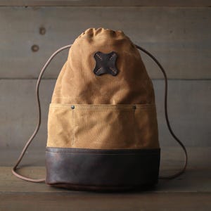 May include: A tan canvas drawstring backpack with a brown leather bottom and a brown leather patch with a cross design. The bag has a drawstring closure and adjustable straps.
