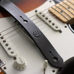 Puede incluir: Primer plano de una correa de guitarra de cuero negro sobre una guitarra el&eacute;ctrica blanca. La correa tiene un logotipo circular y se asegura con un bot&oacute;n. La guitarra tiene pastillas blancas, un mando de volumen y cuerdas.