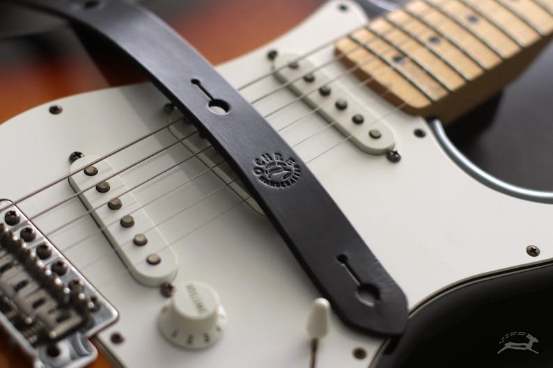 Puede incluir: Primer plano de una correa de guitarra de cuero negro sobre una guitarra el&eacute;ctrica blanca. La correa tiene un logotipo circular y se asegura con un bot&oacute;n. La guitarra tiene pastillas blancas, un mando de volumen y cuerdas.