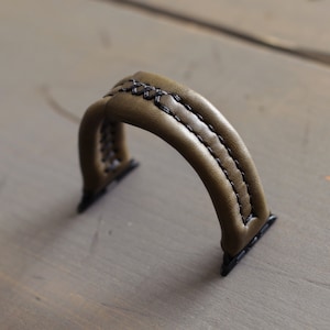 May include: A green leather watch band with black stitching. The band is designed for a smartwatch with a square face.