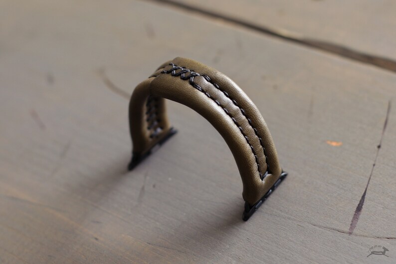 May include: A green leather watch band with black stitching. The band is designed for a smartwatch with a square face.