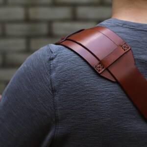 May include: A brown leather shoulder strap with a padded shoulder rest. The strap is attached with decorative stitching. The strap is designed to be worn over the shoulder. The background is a gray brick wall.