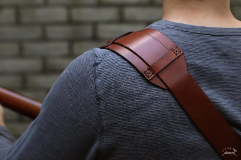 May include: A brown leather shoulder strap with a padded shoulder rest. The strap is attached with decorative stitching. The strap is designed to be worn over the shoulder. The background is a gray brick wall.