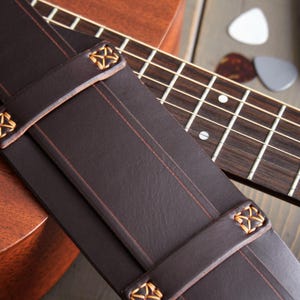 May include: Close-up of a dark brown leather guitar strap with decorative gold stitching and accents. The strap is positioned over a wooden acoustic guitar and fretboard, with guitar picks and a capo visible in the background.