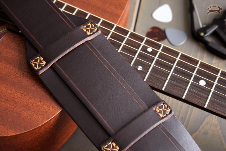 May include: Close-up of a dark brown leather guitar strap with decorative gold stitching and accents. The strap is positioned over a wooden acoustic guitar and fretboard, with guitar picks and a capo visible in the background.