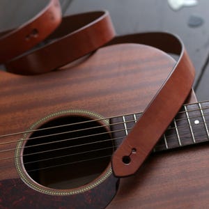 Puede incluir: Guitarra ac&uacute;stica con una correa de cuero marr&oacute;n sobre el cuerpo y el m&aacute;stil. La guitarra tiene un acabado de madera con una boca y cuerdas. Un capotraste plateado es visible en el fondo.
