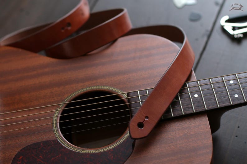 Puede incluir: Guitarra ac&uacute;stica con una correa de cuero marr&oacute;n sobre el cuerpo y el m&aacute;stil. La guitarra tiene un acabado de madera con una boca y cuerdas. Un capotraste plateado es visible en el fondo.