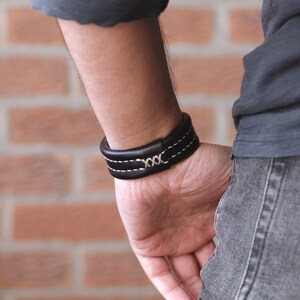 May include: A black leather bracelet with white stitching and a silver 'XXX' detail.