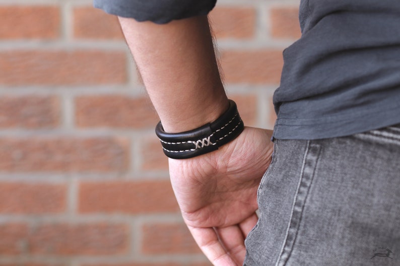 May include: A black leather bracelet with white stitching and a silver 'XXX' detail.