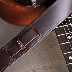 May include: Close-up of a dark brown leather guitar strap with decorative stitching. The strap is positioned over a guitar's fretboard, showcasing the instrument's details. The strap has a buckle and is made of high-quality leather.