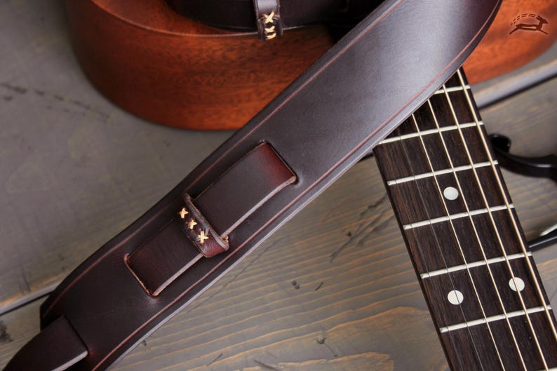 May include: Close-up of a dark brown leather guitar strap with decorative stitching. The strap is positioned over a guitar's fretboard, showcasing the instrument's details. The strap has a buckle and is made of high-quality leather.