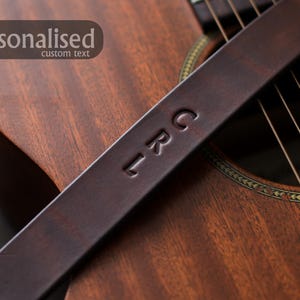 Puede incluir: Una correa de guitarra de cuero marr&oacute;n oscuro con las iniciales "LRC" grabadas en relieve. La correa est&aacute; colocada sobre una guitarra ac&uacute;stica de madera, con la palabra "personalizado" y "texto personalizado" en una pancarta en la esquina superior izquierda.