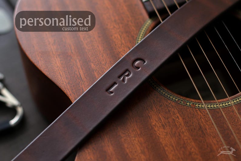 Puede incluir: Una correa de guitarra de cuero marr&oacute;n oscuro con las iniciales "LRC" grabadas en relieve. La correa est&aacute; colocada sobre una guitarra ac&uacute;stica de madera, con la palabra "personalizado" y "texto personalizado" en una pancarta en la esquina superior izquierda.