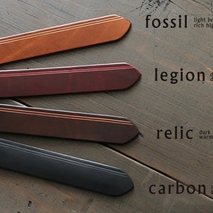Op de afbeelding: Vier leren riemen in verschillende kleuren en texturen, gelabeld "fossil", "legion", "relic" en "carbon" met beschrijvingen van hun kleur en kenmerken.