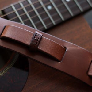 May include: Close-up of a brown leather guitar strap with a stitched detail. The strap is resting on a brown acoustic guitar, with the fretboard and soundhole visible. The strap appears to be made of high-quality leather.