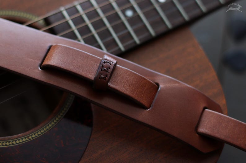 May include: Close-up of a brown leather guitar strap with a stitched detail. The strap is resting on a brown acoustic guitar, with the fretboard and soundhole visible. The strap appears to be made of high-quality leather.