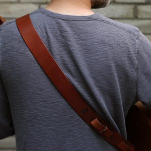 May include: A brown leather guitar strap is draped over the shoulder of a person wearing a gray long-sleeve shirt. The strap is attached to a brown acoustic guitar. The background is a brick wall.