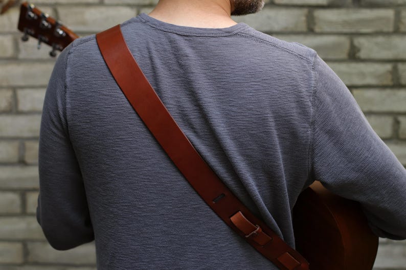 May include: A brown leather guitar strap is draped over the shoulder of a person wearing a gray long-sleeve shirt. The strap is attached to a brown acoustic guitar. The background is a brick wall.