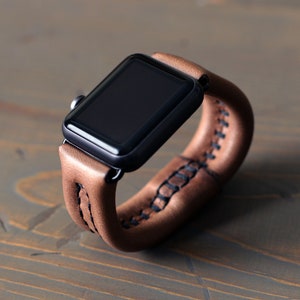 May include: A brown leather watch band with a black Apple Watch. The band has a double wrap design and is stitched with black thread.