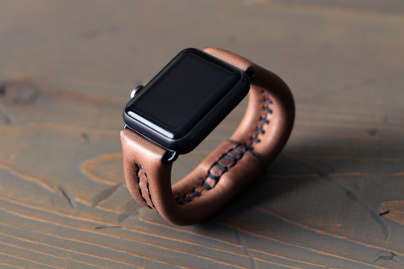 May include: A brown leather watch band with a black Apple Watch. The band has a double wrap design and is stitched with black thread.