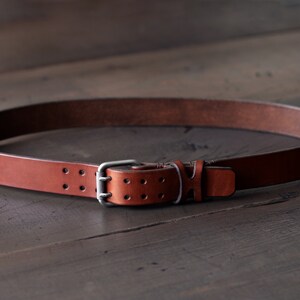 May include: A brown leather belt with a silver buckle and double holes. The belt is lying on a wooden surface.
