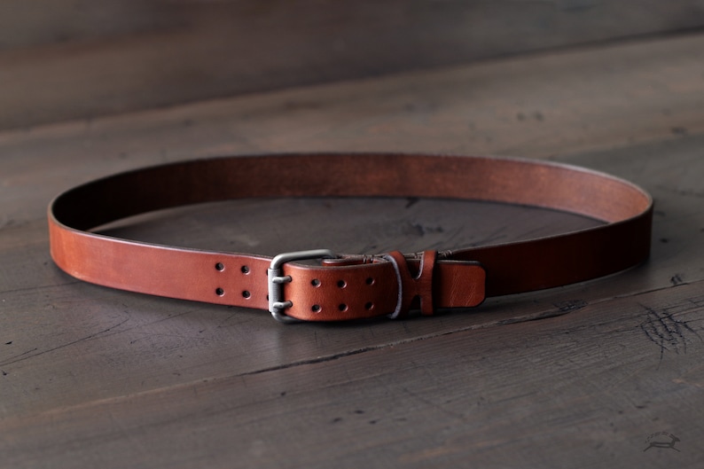 May include: A brown leather belt with a silver buckle and double holes. The belt is lying on a wooden surface.