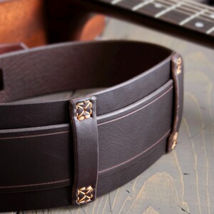 May include: Close-up of a dark brown leather guitar strap with decorative stitching and accents. The strap has a classic design with a subtle texture and is designed to be worn over the shoulder. A guitar is in the background.