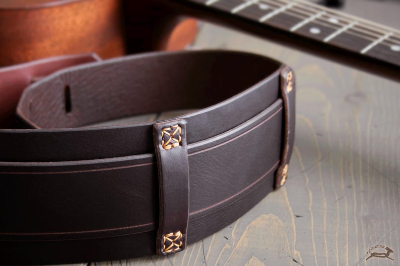 May include: Close-up of a dark brown leather guitar strap with decorative stitching and accents. The strap has a classic design with a subtle texture and is designed to be worn over the shoulder. A guitar is in the background.