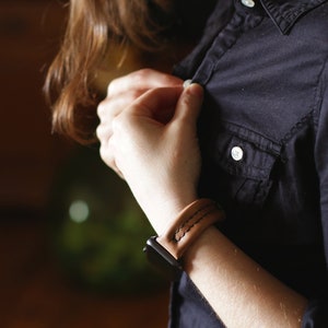 May include: A brown leather watch band with white stitching. The band is worn on a person's wrist and is fastened with a black buckle.