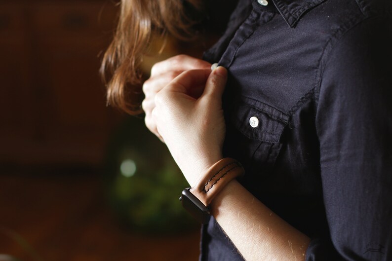 May include: A brown leather watch band with white stitching. The band is worn on a person's wrist and is fastened with a black buckle.