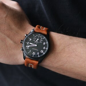 May include: A black smartwatch with a brown leather band. The watch face has a black background with white numbers and hands. The watch has a chronograph feature.