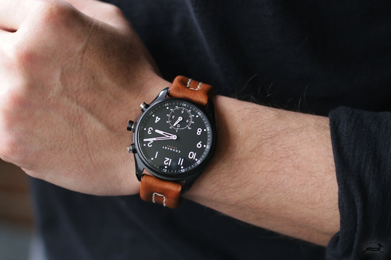 May include: A black smartwatch with a brown leather band. The watch face has a black background with white numbers and hands. The watch has a chronograph feature.