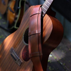 May include: Acoustic guitar with a brown leather strap. The guitar has a wooden body with a sound hole and a red pickguard. The strap is attached to the guitar's body with decorative stitching. The guitar is resting on a wooden surface.