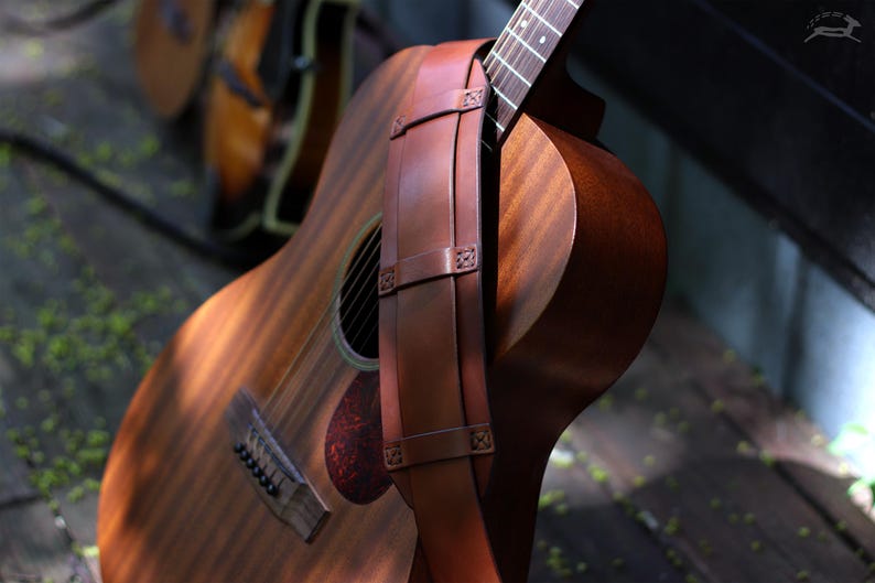 May include: Acoustic guitar with a brown leather strap. The guitar has a wooden body with a sound hole and a red pickguard. The strap is attached to the guitar's body with decorative stitching. The guitar is resting on a wooden surface.