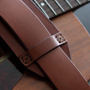 May include: Close-up of a brown leather guitar strap with decorative stitching. The strap is draped over a wooden acoustic guitar, showcasing the instrument's sound hole and fretboard. The strap's rich color complements the guitar's wood grain.