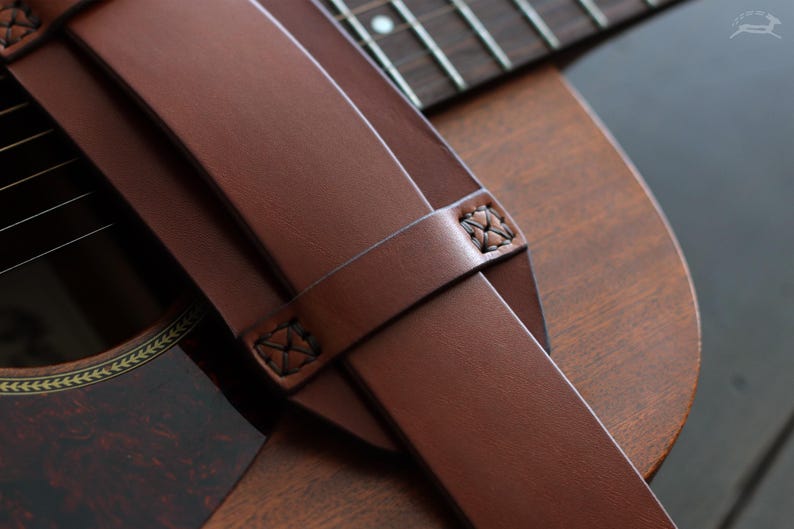 May include: Close-up of a brown leather guitar strap with decorative stitching. The strap is draped over a wooden acoustic guitar, showcasing the instrument's sound hole and fretboard. The strap's rich color complements the guitar's wood grain.