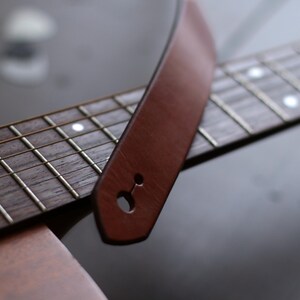 Puede incluir: Primer plano de una correa de guitarra de cuero marr&oacute;n sobre un diapas&oacute;n de guitarra. La correa tiene un solo agujero para la fijaci&oacute;n. El diapas&oacute;n es de madera oscura con trastes plateados e incrustaciones blancas.