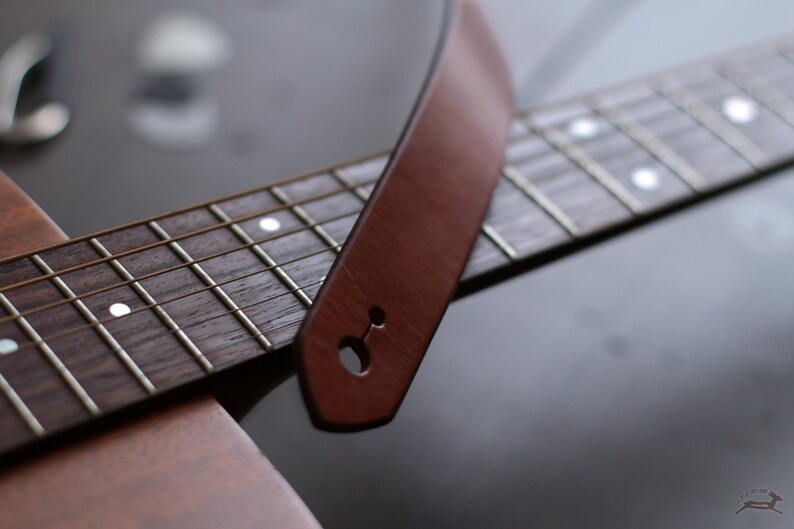 Puede incluir: Primer plano de una correa de guitarra de cuero marr&oacute;n sobre un diapas&oacute;n de guitarra. La correa tiene un solo agujero para la fijaci&oacute;n. El diapas&oacute;n es de madera oscura con trastes plateados e incrustaciones blancas.