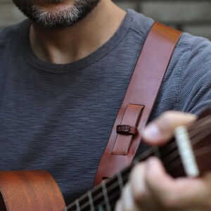 May include: A brown leather guitar strap with a shoulder pad is shown. The strap is attached to a brown acoustic guitar. The person wearing a gray long-sleeve shirt is playing the guitar.