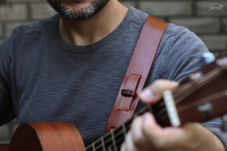 May include: A brown leather guitar strap with a shoulder pad is shown. The strap is attached to a brown acoustic guitar. The person wearing a gray long-sleeve shirt is playing the guitar.