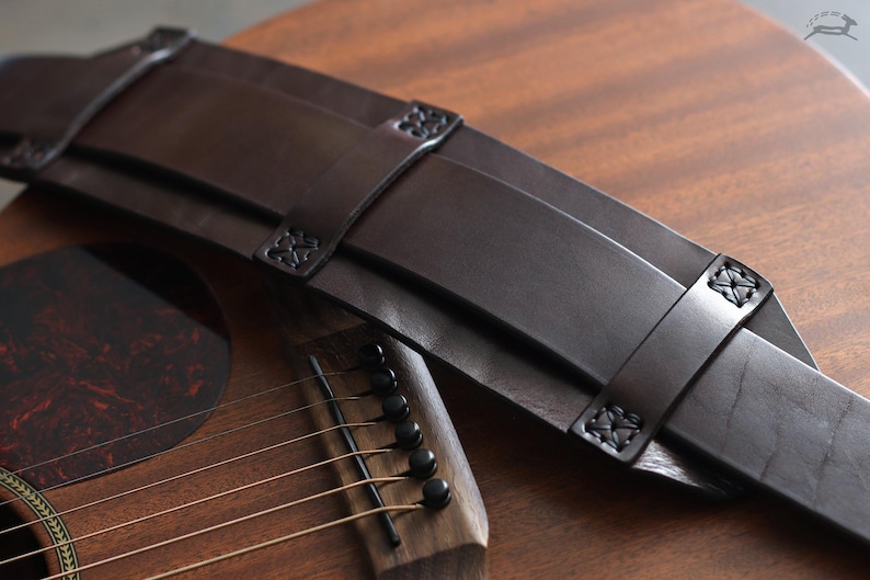 May include: A dark brown leather guitar strap with decorative stitching and embossed details. The strap is shown resting on a wooden acoustic guitar, showcasing its rich color and texture. The strap's design suggests a focus on both aesthetics and functionality.