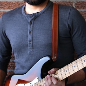 Puede incluir: Una guitarra el&eacute;ctrica marr&oacute;n y negra con m&aacute;stil de madera clara es sostenida por una persona que lleva una camisa gris de manga larga. Se ve una correa de guitarra de cuero marr&oacute;n. El fondo es una pared de ladrillo.