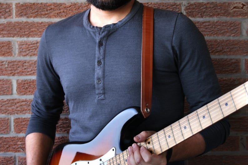Puede incluir: Una guitarra el&eacute;ctrica marr&oacute;n y negra con m&aacute;stil de madera clara es sostenida por una persona que lleva una camisa gris de manga larga. Se ve una correa de guitarra de cuero marr&oacute;n. El fondo es una pared de ladrillo.