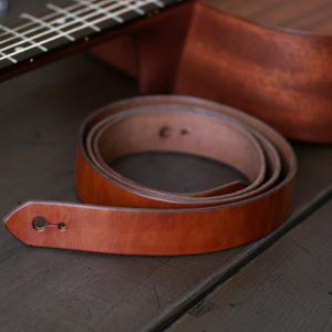 Puede incluir: Una correa de guitarra enrollada de cuero marr&oacute;n claro con un ojal en el extremo. Una ukelele marr&oacute;n est&aacute; en el fondo. El conjunto est&aacute; sobre una superficie de madera oscura.