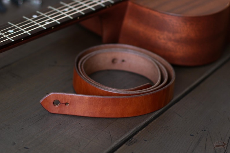 Puede incluir: Una correa de guitarra enrollada de cuero marr&oacute;n claro con un ojal en el extremo. Una ukelele marr&oacute;n est&aacute; en el fondo. El conjunto est&aacute; sobre una superficie de madera oscura.