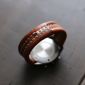 May include: A brown leather watch strap with white stitching. The strap is folded in half and the watch face is visible.