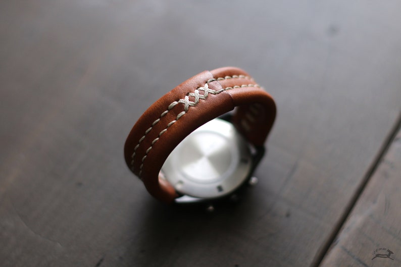 May include: A brown leather watch strap with white stitching. The strap is folded in half and the watch face is visible.