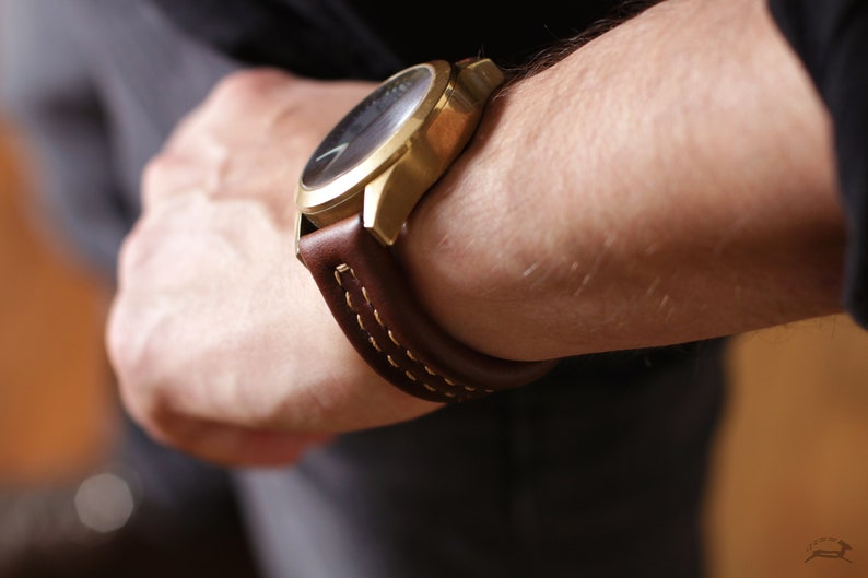 May include: A gold-tone wristwatch with a brown leather band and white stitching. The watch has a black face.