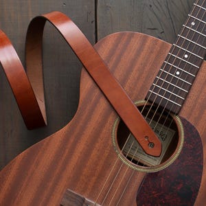 Puede incluir: Guitarra ac&uacute;stica con una correa de cuero marr&oacute;n. La guitarra tiene un cuerpo de madera con un golpeador marr&oacute;n oscuro y una boca. La correa est&aacute; enganchada a la guitarra y es de un color marr&oacute;n c&aacute;lido y rico.